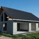 Budynek w stanie surowym otwartym, z betonowych pustaków, z dachem pokrytym czarną dachówką. Widać otwory okienne i drzwiowe.