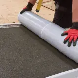 Ręce w rękawiczkach rozwijają papę podkładową na dachu.