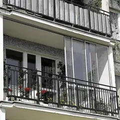 Zabudowa balkonu w bloku z przeszklonymi drzwiami i metalową balustradą. Na balkonie stoją doniczki z kwiatami.