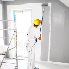 Malowanie pokoju na szaro. Pracownik w żółtym kasku maluje ścianę wałkiem. Zastanawiasz się, ile kosztuje malowanie pokoju?