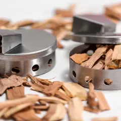 Dwa metalowe pojemniki wypełnione wiórami drzewnymi do wędzenia.