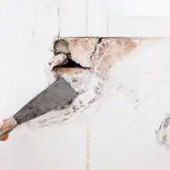 Dłonią z kielnią usuwa się luźny tynk, przygotowując ścianę do wypełnienia dużych ubytków w ścianie wewnętrznej.