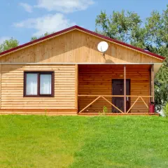Drewniany domek z werandą, otoczony zielenią. Może to być dom bez pozwolenia, ale wygląda przytulnie.