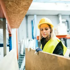 Kobieta w kasku i kamizelce odblaskowej przygotowuje płytę OSB, być może do projektu, jak zrobić drzwi z płyty OSB.