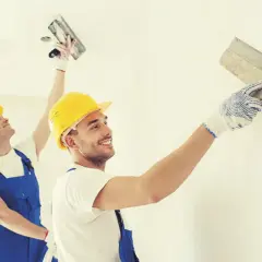 Dwóch mężczyzn w żółtych kaskach i niebieskich ogrodniczkach nakłada tynk na ścianę. Różne rodzaje tynków wewnętrznych to ich specjalność.