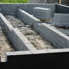 Budowa fundamentów z bloczków betonowych. Widać postęp prac nad izolacją pionową fundamentów, ułożone palety z materiałem budowlanym.