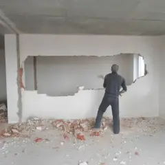 Mężczyzna z młotem burzy ścianę nośną, tworząc otwór. Gruz leży na podłodze.