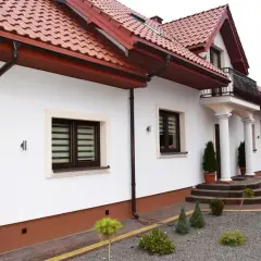 Biały dom z brązowym dachem i rynnami. Elegancki cokół podkreśla jego architekturę.