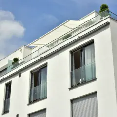 Nowoczesny budynek z białym tynkiem, przeszklonymi balustradami i markizą. Widać kilka balkonów francuskich z roślinnością.