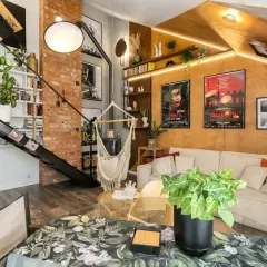 Salon w stylu loftowym z ceglaną ścianą, metalowymi schodami, wiszącym fotelem i wygodną kanapą.