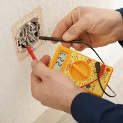 Elektryk sprawdza napięcie w gniazdku za pomocą żółtego multimetru. Wynik pomiaru to 221V.