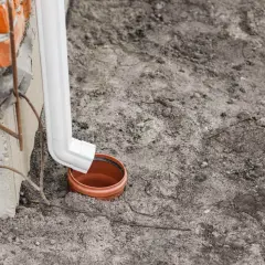 Biała rura spustowa odprowadza wodę z rynien do gruntu, do ceramicznej studzienki.