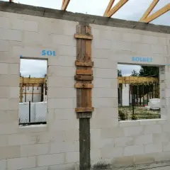 Ściana z bloczków betonowych z otworami okiennymi i drzwiami. Widoczny betonowy nadproże nad drzwiami, drewniane szalunki i elementy konstrukcji dachu.