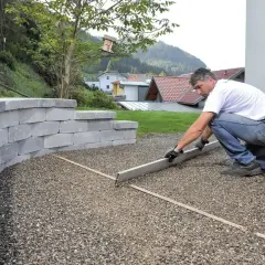 Mężczyzna wyrównuje żwir na nowym podjeździe z kamienia. W tle widać domy i wzgórza.