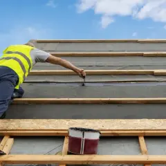 Budowlaniec w kamizelce zakłada papę na dach z desek. Podkład dachowy i drewniane łaty tworzą solidną konstrukcję.