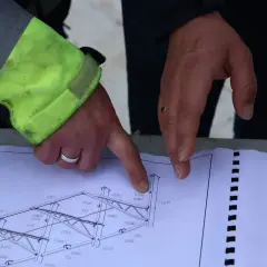 Rysunek techniczny konstrukcji stalowej, omawiany podczas zgłoszenia budowy. Dłonie wskazują na szczegóły projektu.
