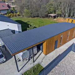 Nowoczesny budynek z drewnianą elewacją i ciemnym, **dachem jednospadowym**. Obok stoi samochód i przyczepa pod wiatą.