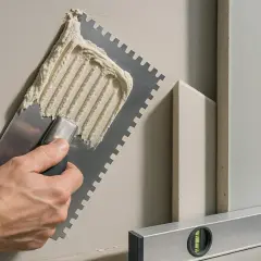 Ręka z kielnią rozprowadza klej do płyt gk. Obok leżą płyty i poziomica.