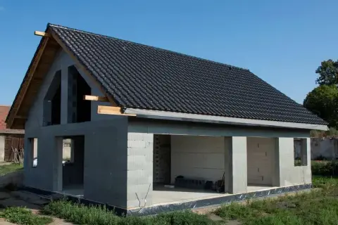 Budynek w stanie surowym otwartym, z betonowych pustaków, z dachem pokrytym czarną dachówką. Widać otwory okienne i drzwiowe.