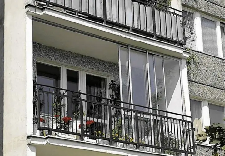 Zabudowa balkonu w bloku z przeszklonymi drzwiami i metalową balustradą. Na balkonie stoją doniczki z kwiatami.