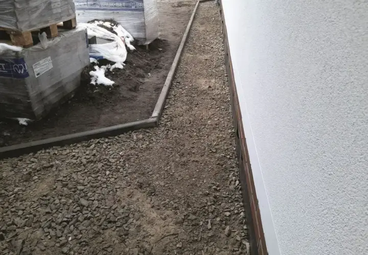 Wykonano opaskę wokół domu z żwiru, obok ułożone są palety z materiałami budowlanymi.