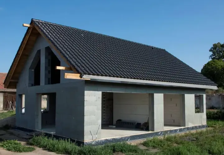 Budynek w stanie surowym otwartym, z betonowych pustaków, z dachem pokrytym czarną dachówką. Widać otwory okienne i drzwiowe.