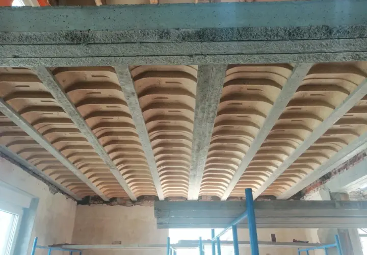Widok od dołu na betonowe belki stropowe i pustaki ceramiczne, tworzące przestrzeń przypominającą sufit katedralny.