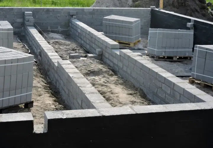Budowa fundamentów z bloczków betonowych. Widać postęp prac nad izolacją pionową fundamentów, ułożone palety z materiałem budowlanym.