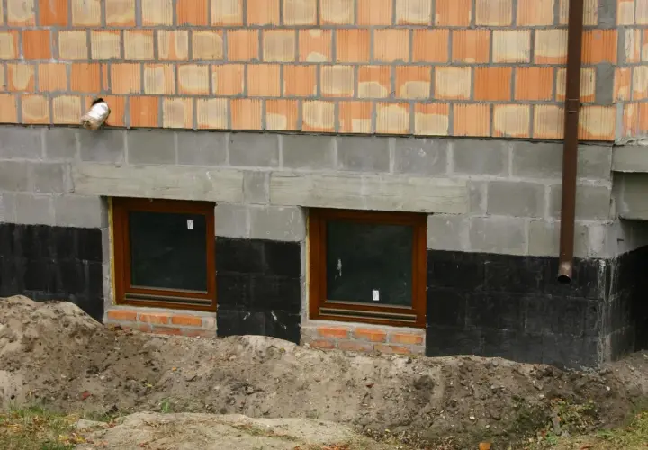 Izolacja fundamentów w starym domu: czarna papa chroni mury przed wilgocią, widać dwa okna piwniczne i ceglaną ścianę.