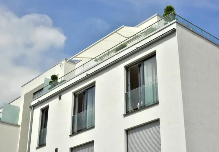 Nowoczesny budynek z białym tynkiem, przeszklonymi balustradami i markizą. Widać kilka balkonów francuskich z roślinnością.
