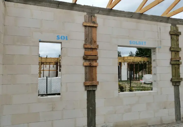 Ściana z bloczków betonowych z otworami okiennymi i drzwiami. Widoczny betonowy nadproże nad drzwiami, drewniane szalunki i elementy konstrukcji dachu.