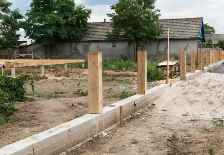 Solidny fundament pod ogrodzenie z drewnianymi słupkami, w tle budowa domu.