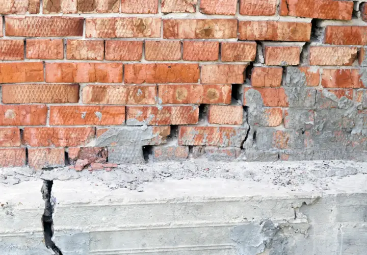 Uszkodzona ceglana ściana z widocznym pęknięciem w betonie, wskazującym na podbijanie fundamentów.