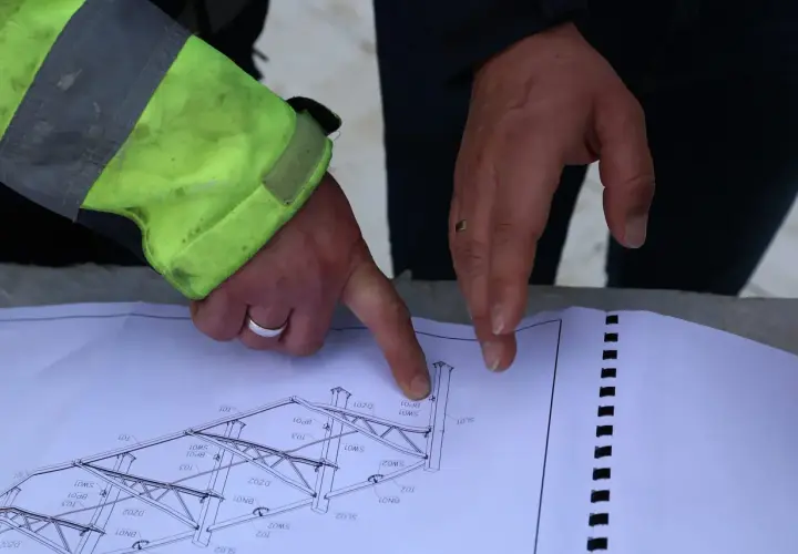 Rysunek techniczny konstrukcji stalowej, omawiany podczas zgłoszenia budowy. Dłonie wskazują na szczegóły projektu.