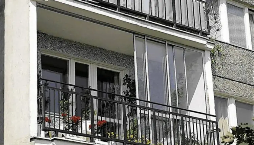 Zabudowa balkonu w bloku z przeszklonymi drzwiami i metalową balustradą. Na balkonie stoją doniczki z kwiatami.