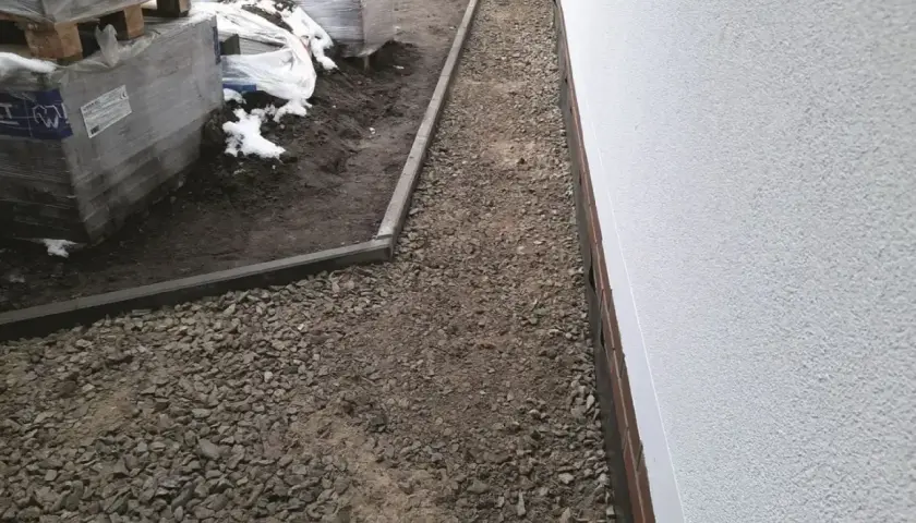 Wykonano opaskę wokół domu z żwiru, obok ułożone są palety z materiałami budowlanymi.