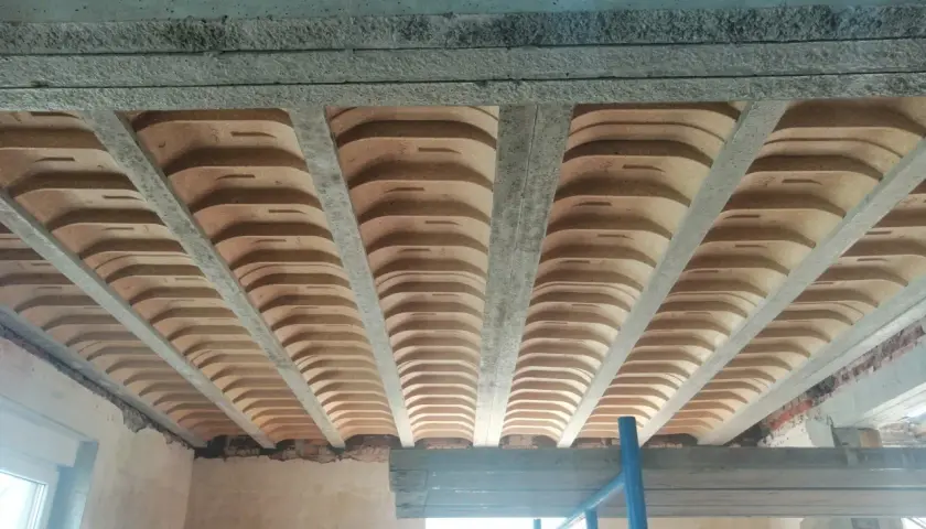 Widok od dołu na betonowe belki stropowe i pustaki ceramiczne, tworzące przestrzeń przypominającą sufit katedralny.