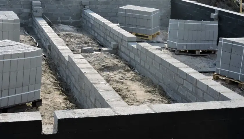 Budowa fundamentów z bloczków betonowych. Widać postęp prac nad izolacją pionową fundamentów, ułożone palety z materiałem budowlanym.