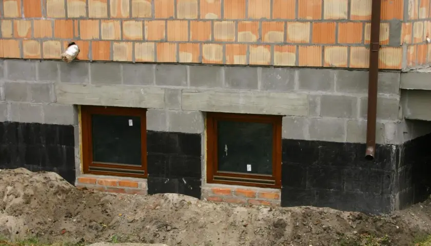 Izolacja fundamentów w starym domu: czarna papa chroni mury przed wilgocią, widać dwa okna piwniczne i ceglaną ścianę.