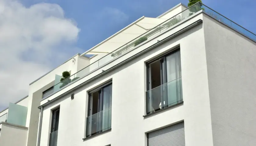 Nowoczesny budynek z białym tynkiem, przeszklonymi balustradami i markizą. Widać kilka balkonów francuskich z roślinnością.