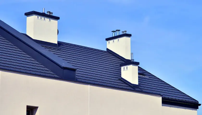 Nowoczesny dach bezokapowy z ciemnogranatową dachówką i białymi kominami, pod błękitnym niebem.