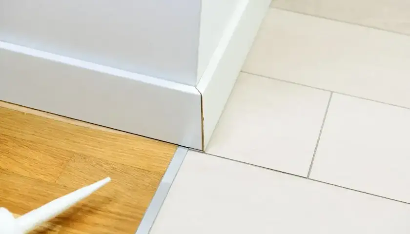 Biała listwa przypodłogowa łączy drewnianą podłogę z płytkami. Widać pistolet z masą uszczelniającą, gotową do wypełnienia szczeliny jako korek dylatacyjny.