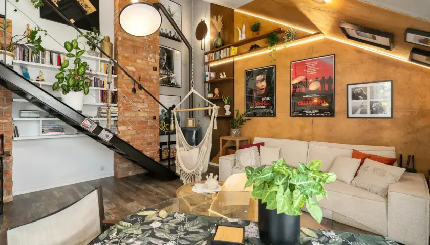 Salon w stylu loftowym z ceglaną ścianą, metalowymi schodami, wiszącym fotelem i wygodną kanapą.