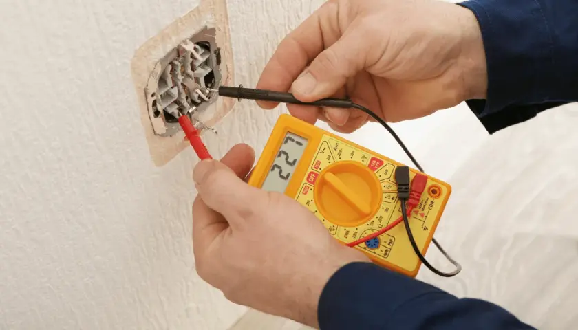 Elektryk sprawdza napięcie w gniazdku za pomocą żółtego multimetru. Wynik pomiaru to 221V.