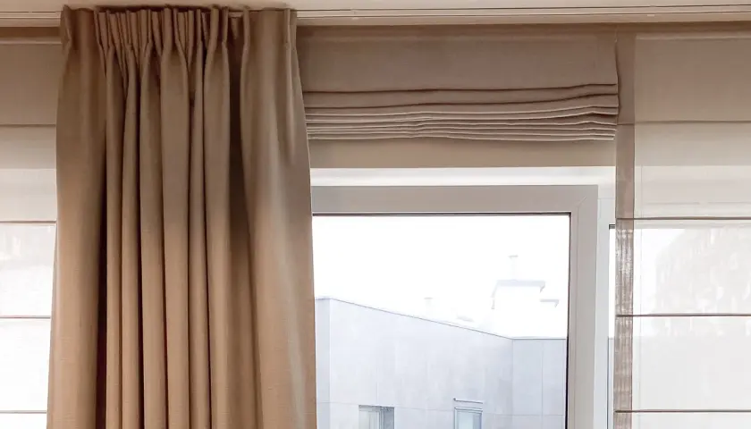 Beżowa roleta rzymska na okno dachowe, częściowo zasłonięta przez zasłonę.