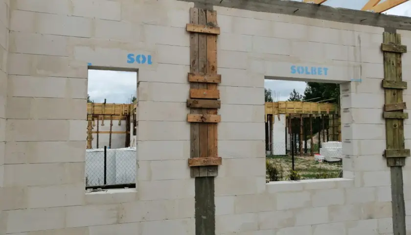 Ściana z bloczków betonowych z otworami okiennymi i drzwiami. Widoczny betonowy nadproże nad drzwiami, drewniane szalunki i elementy konstrukcji dachu.
