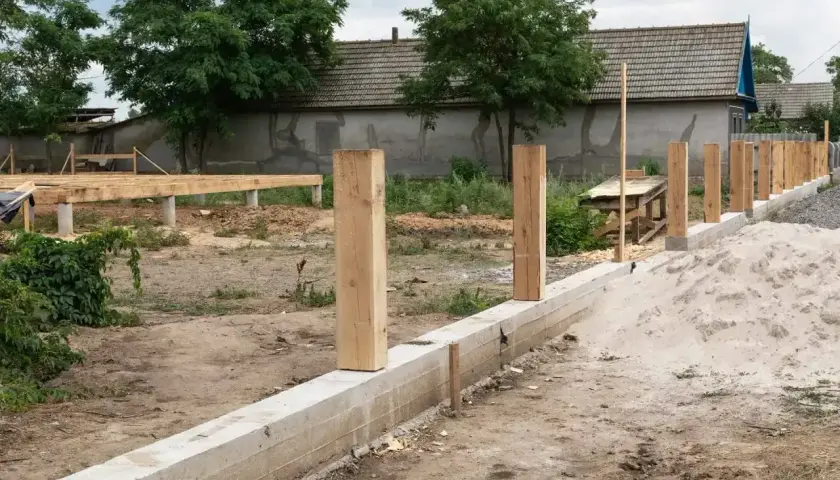 Solidny fundament pod ogrodzenie z drewnianymi słupkami, w tle budowa domu.