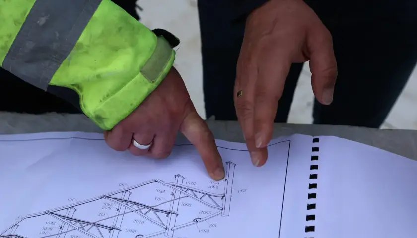 Rysunek techniczny konstrukcji stalowej, omawiany podczas zgłoszenia budowy. Dłonie wskazują na szczegóły projektu.