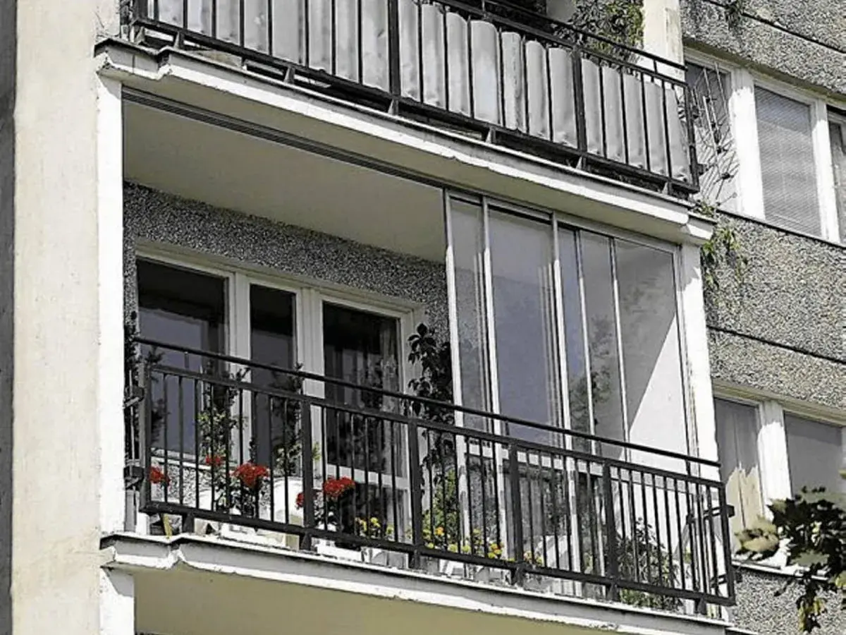 Zabudowa balkonu w bloku z przeszklonymi drzwiami i metalową balustradą. Na balkonie stoją doniczki z kwiatami.