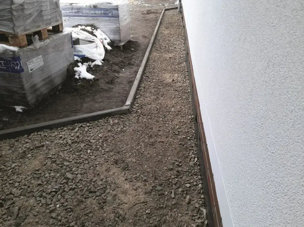 Wykonano opaskę wokół domu z żwiru, obok ułożone są palety z materiałami budowlanymi.